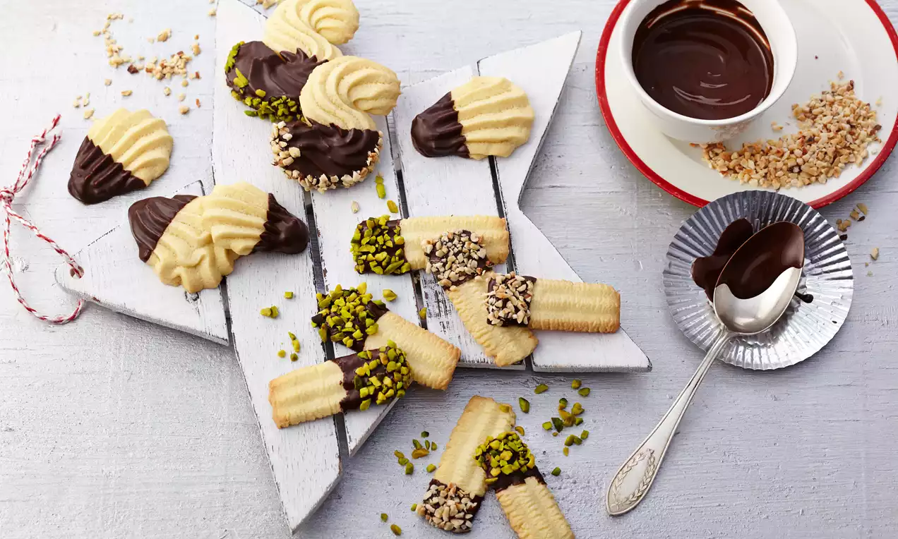 Spritzgebäck in hellgoldener Farbe, teils mit dunkler Schokolade überzogen und mit Pistazien oder gehackten Nüssen dekoriert auf weißem Holzbrett