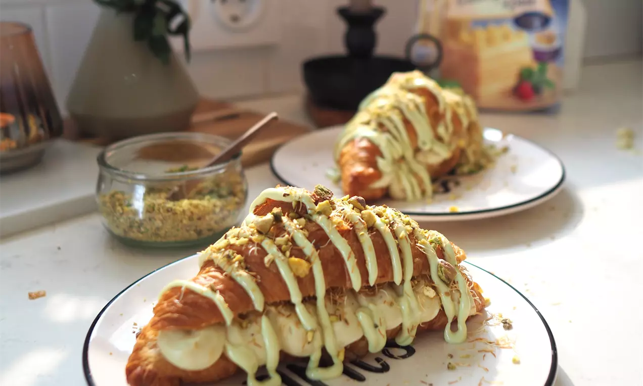 Златисти кроасани с хрупкава текстура, декорирани с крем от шамфъстък и натрошени ядки върху бяла чиния в пастелна обстановка