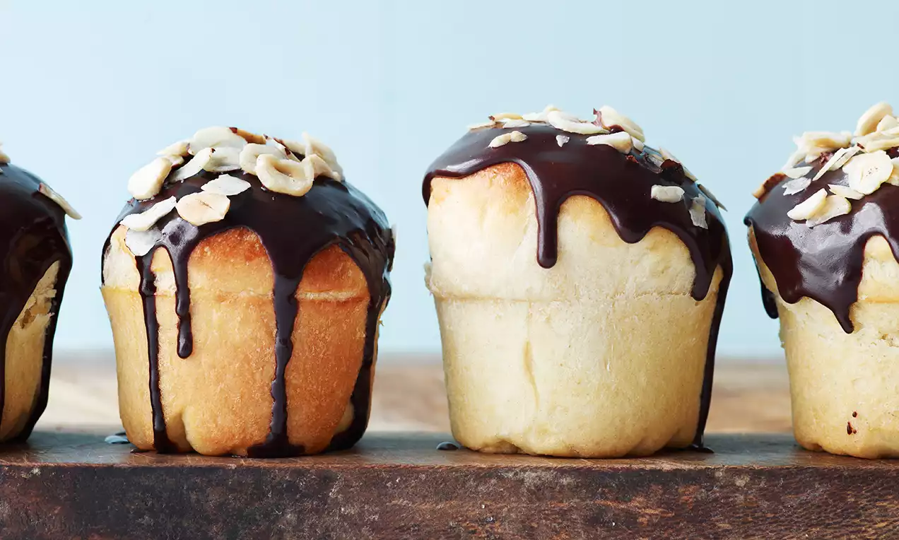 Gyllenbrune muffinboller med blank sjokoladekrem som drypper langs kantene, toppet med lyse mandelflak, stående på mørkt treunderlag
