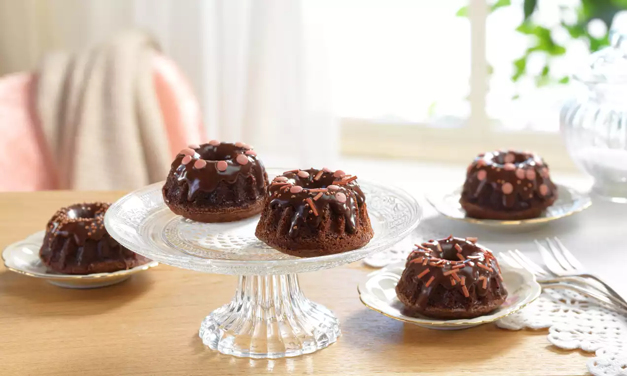 Mini-Schoko-Gugelhupfe mit glänzender Schokoglasur und rosa Streuseln auf einer gläsernen Tortenplatte und zarten Porzellantellern angerichtet