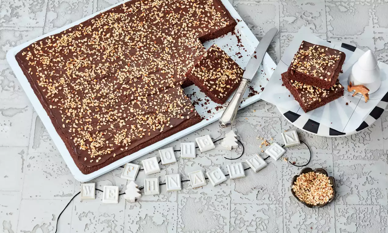 Braunes Schokoladenbrot mit glänzender Oberfläche und gehackten Nüssen, serviert neben einem Teller mit gestapelten Stücken und Weihnachtsdekor