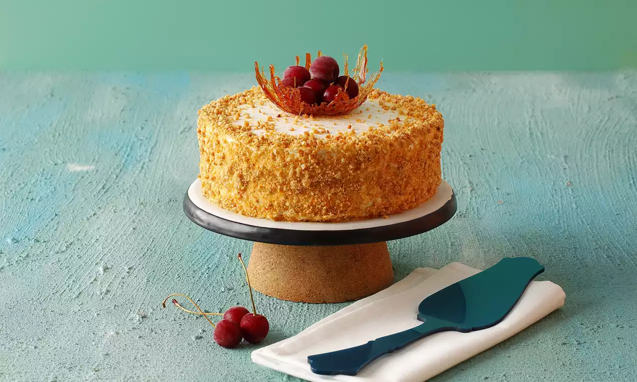 Altın krokan kaplı yuvarlak pasta, karamelize dekor ve parlak kırmızı kirazlarla süslenerek ayaklı bir tabağa yerleştirilmiş