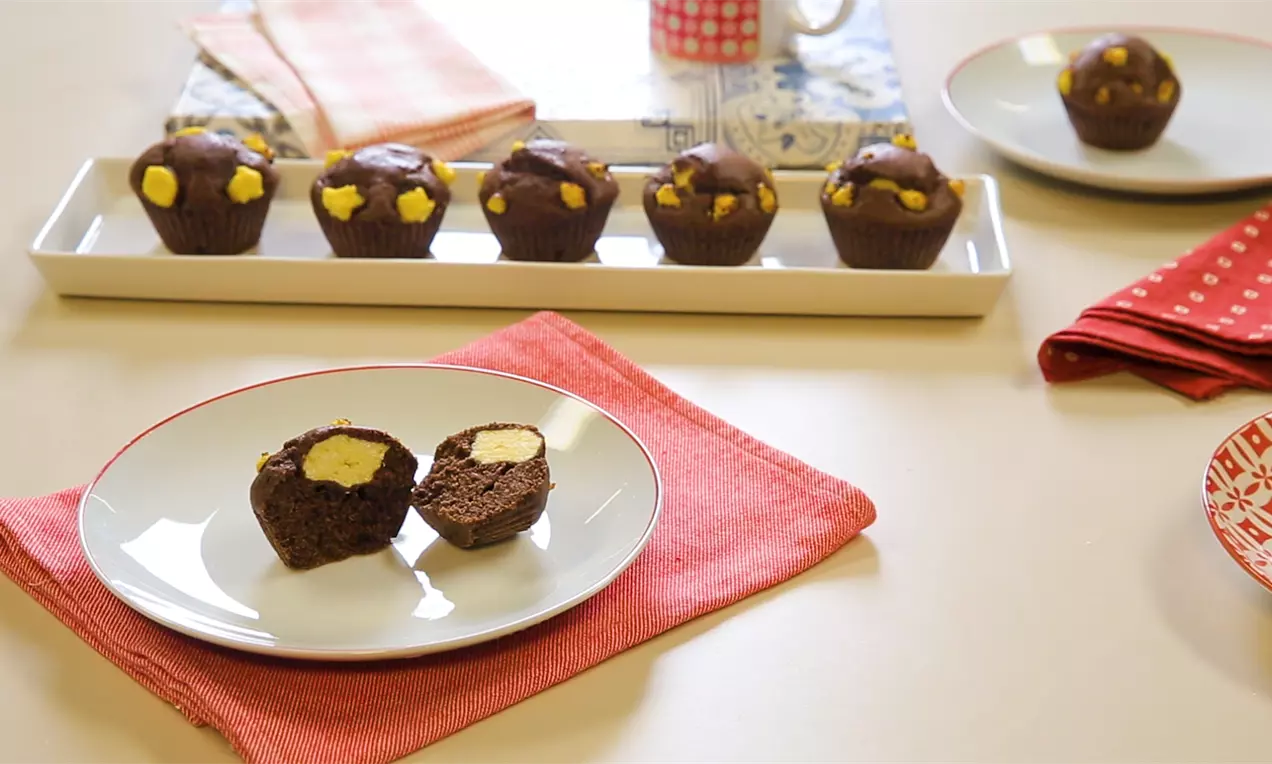 Шоколадови мъфини с ярко бели парченца крем, подредени върху правоъгълна бяла чиния, с разрязани мъфини на преден план върху червен плат