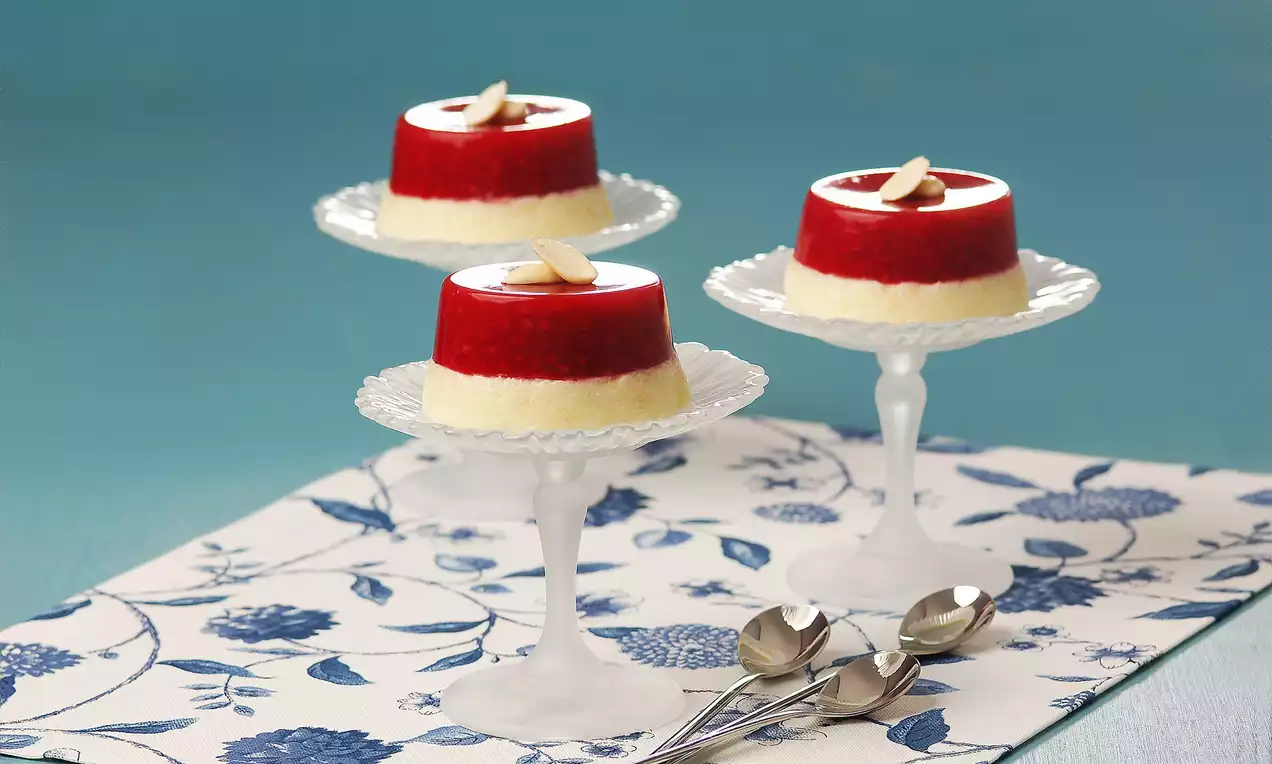 Kırmızı ahududu soslu, krem rengi irmik tatlısı katmanlı, küçük cam ayaklı tabaklarda badem dilimiyle süslenmiş şekilde sunulmuş