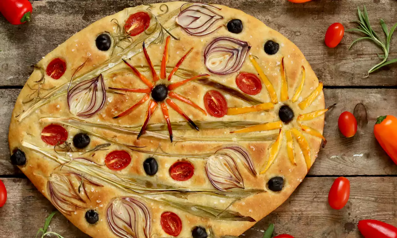 Oval focaccia med gyllen skorpe dekorert med røde tomater, svarte oliven, løkstrimler og grønne urter plassert på rustikt trebord