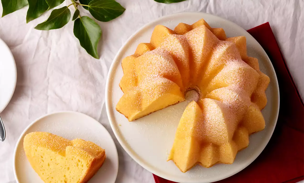 Goldgelber Topfengugelhupf mit feinen Zuckerkristallen, angeschnitten auf weißem Teller, daneben ein Kuchenstück und grüne Blätter