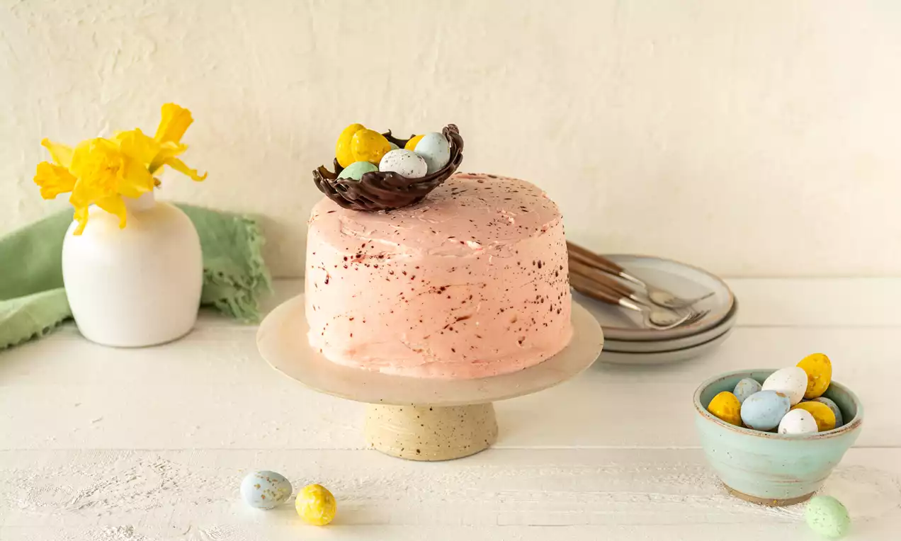 Gâteau rose pâle aux éclats de chocolat, décoré d’un nid en chocolat brun avec des œufs colorés, posé sur un présentoir beige