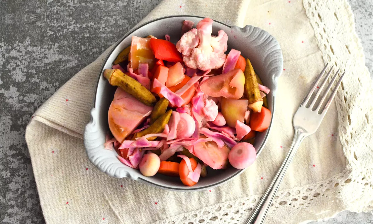 Пъстра смес от розовосиви резени бамя, лилаво зеле, зелени краставички и бели гъби, подредени в бяла купа върху ленена салфетка