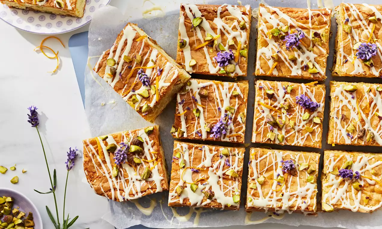 Golden blondie squares topped with drizzled white icing, chopped pistachios, orange zest, and purple lavender sprigs on parchment paper