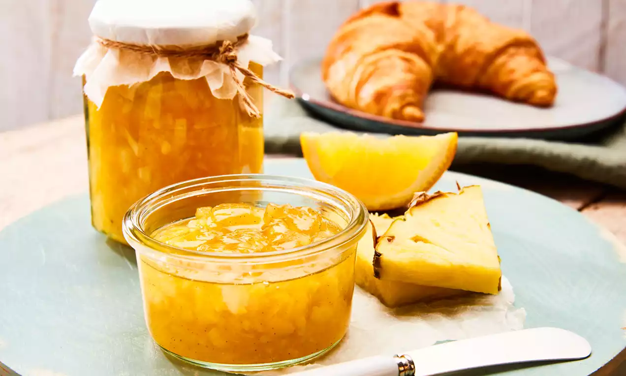 Ananas-Orangenmarmelade mit Bourbon-Vanille im Glas, glänzend goldorange mit Vanillesamen und Fruchtstücken, dazu Orangenscheiben und Löffel