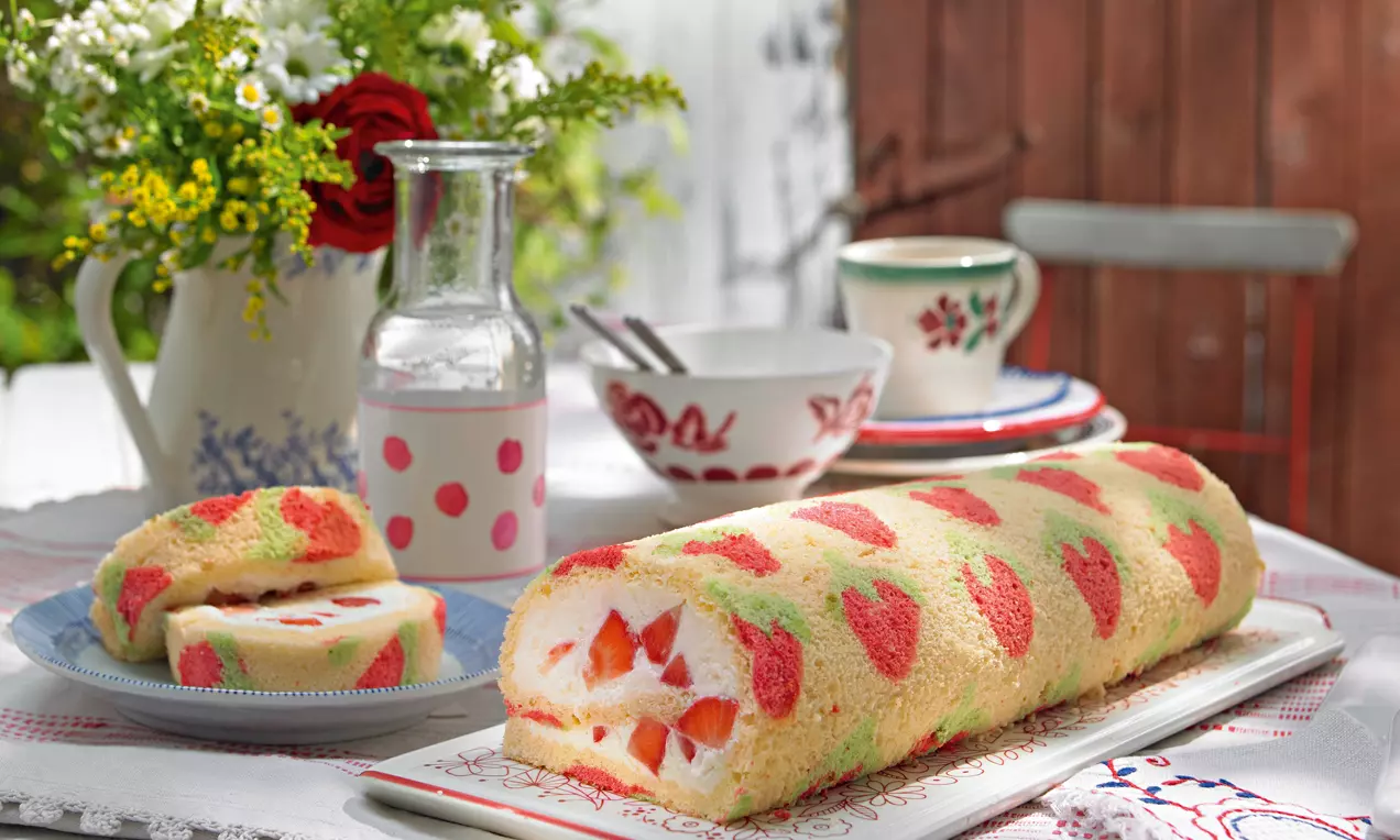 Svijetla rolada s uzorkom crvenih jagoda, s bijelim kremastim punjenjem i komadićima jagoda, poslužena na tanjuru na stolu