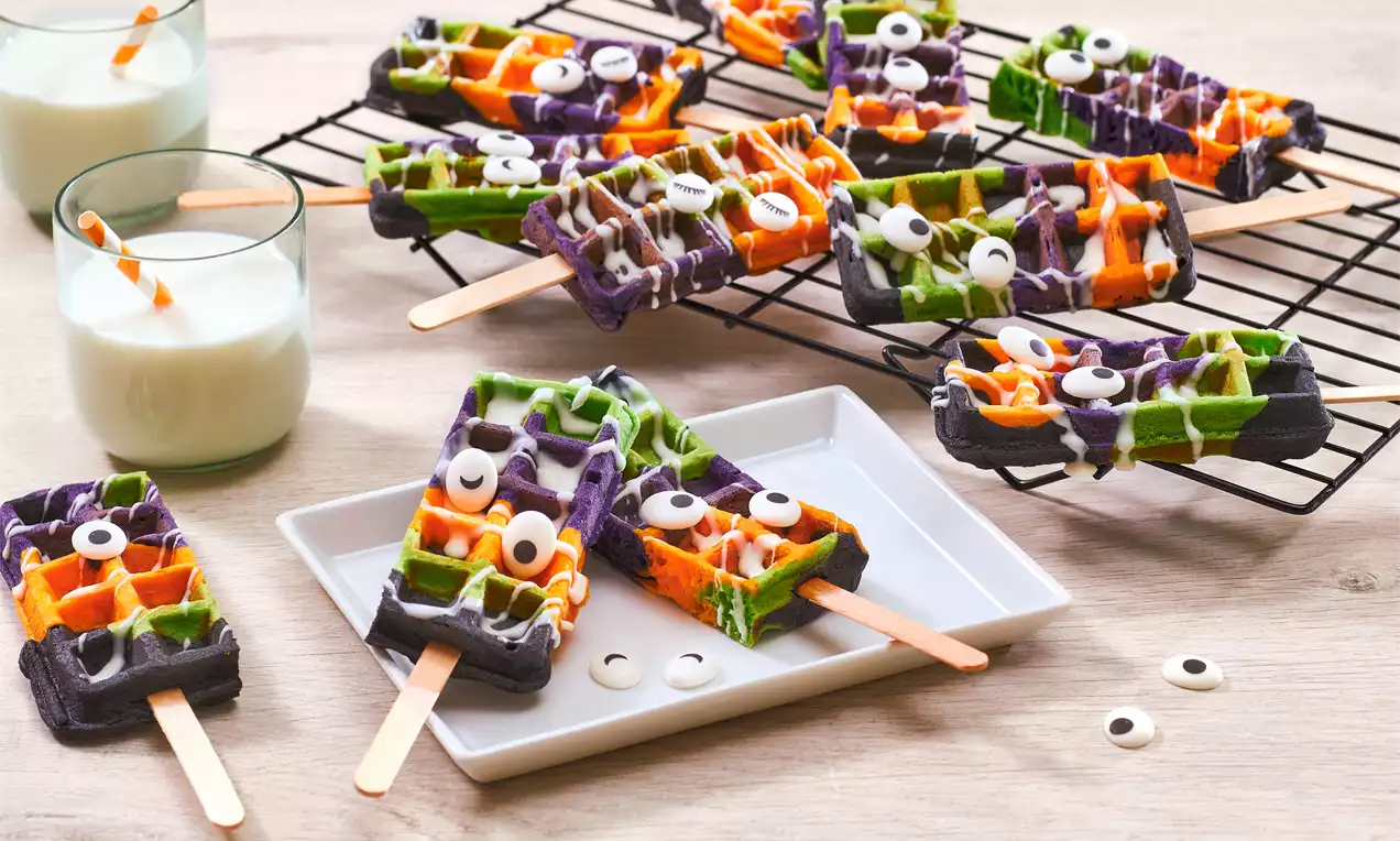Gaufres multicolores sur bâtonnets en nuances de vert, orange, violet et noir, décorées d'yeux en sucre et filet de chocolat blanc
