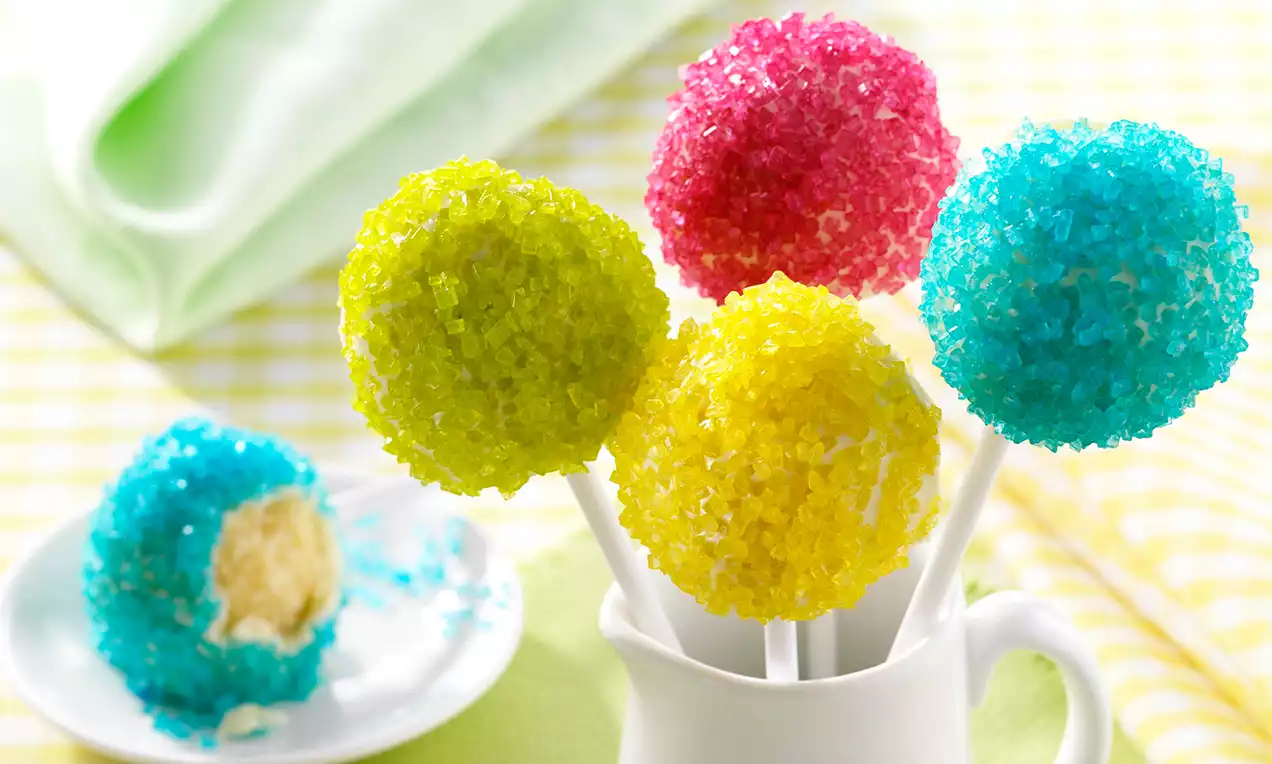 Värikkäitä pyöreitä kakkutikkareita, päällinen kimaltelevista sokerikiteistä, aseteltu valkoiseen mukiin kirkkaassa pöytäympäristössä