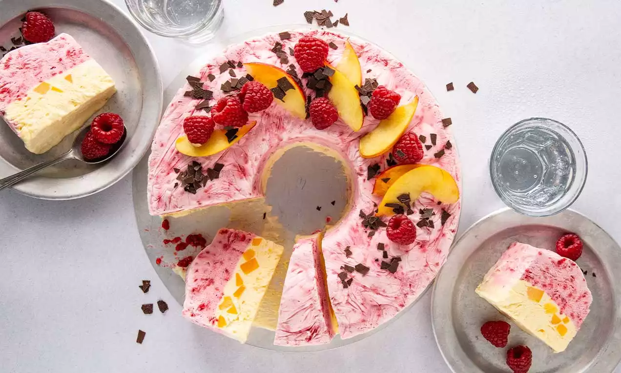 Ringförmiges Himbeer-Pfirsich-Parfait mit rosa-weißem Muster, frischen Himbeeren, gelben Pfirsichspalten und Schokoraspeln auf einem Teller