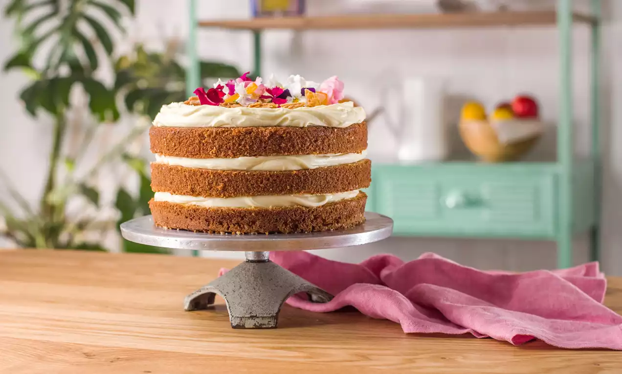 Pastel de capas doradas con crema blanca, decorado con flores y piel de naranja sobre un soporte metálico en una cocina luminosa