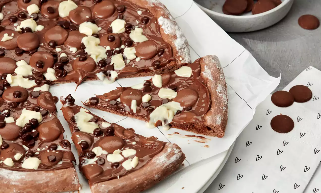 Runde Schoko-Pizza mit glänzendem Belag aus dunkler Creme, weißen und braunen Schokoladenstücken auf weißem Backpapier mit einer Schale Schokoladenplättchen im Hintergrund