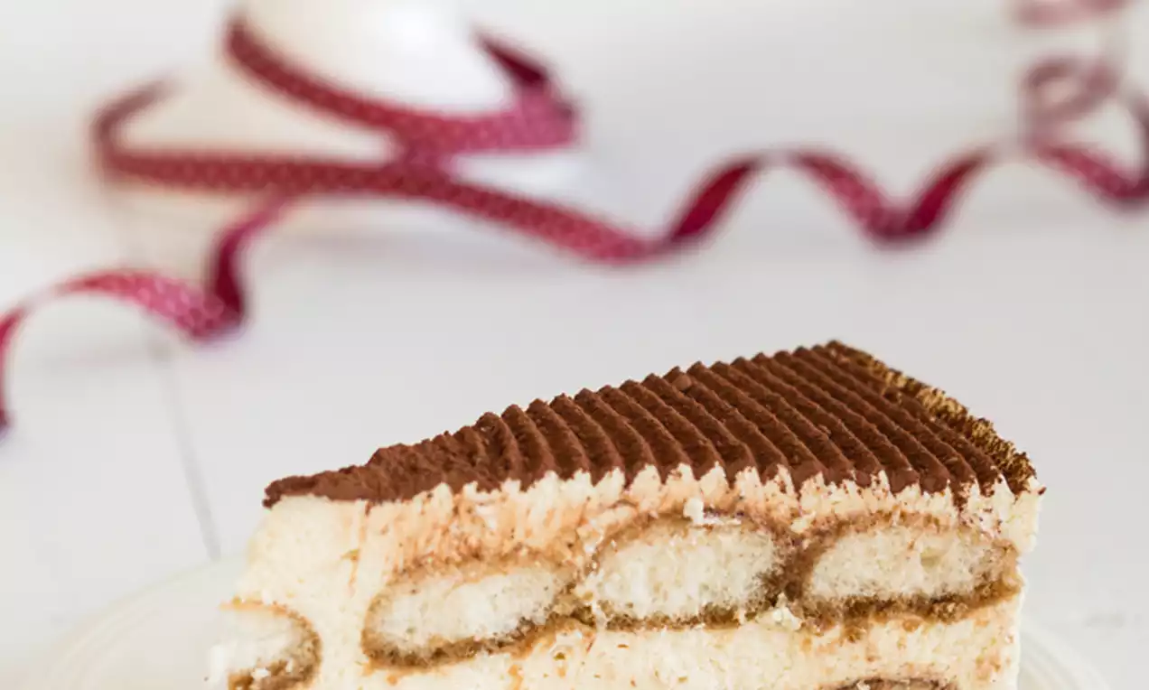 Kawałek tiramisu na białym talerzu, z jasnokremowym nadzieniem, biszkoptami nasączonymi kawą, ciemnym kakaowym wierzchem i srebrną łyżeczką obok