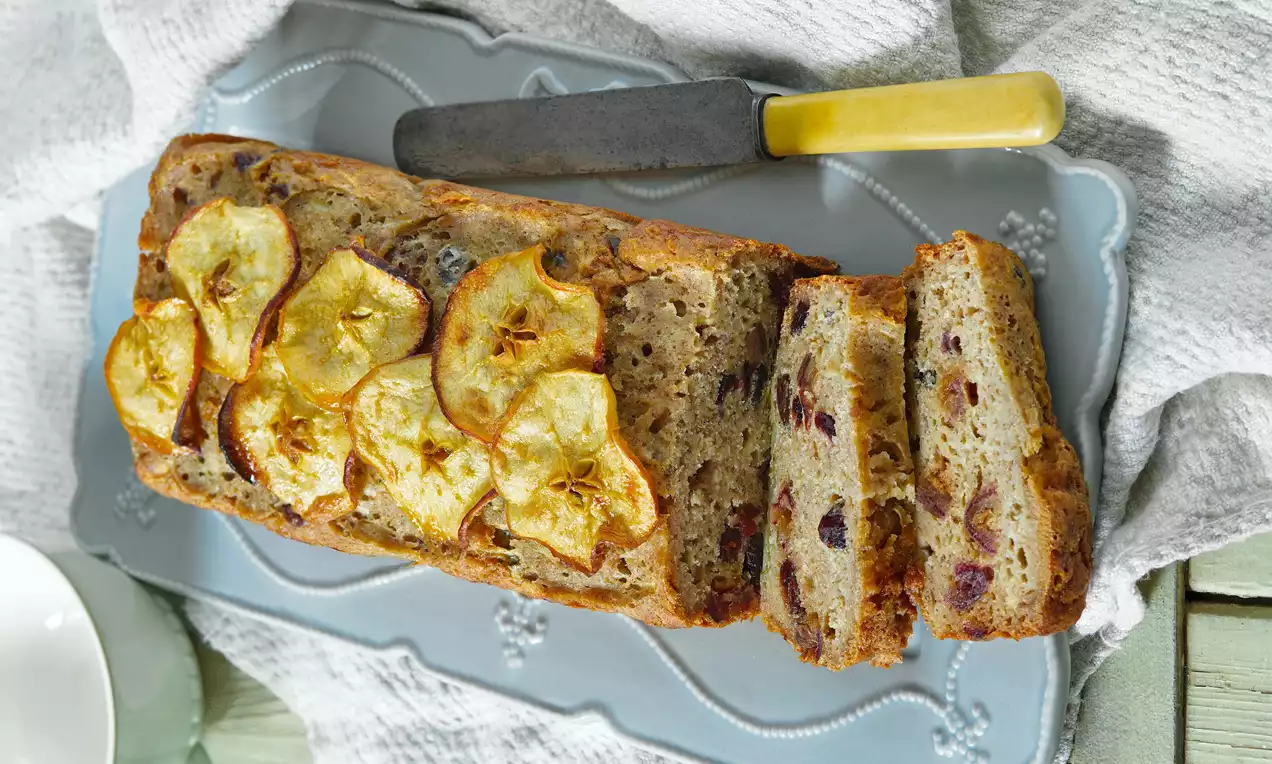 Podlouhlý jablečný chlebíček zlatavé barvy s kousky brusinek a plátky pečených jablek na zdobeném modrém talíři vedle nože s žlutou rukojetí