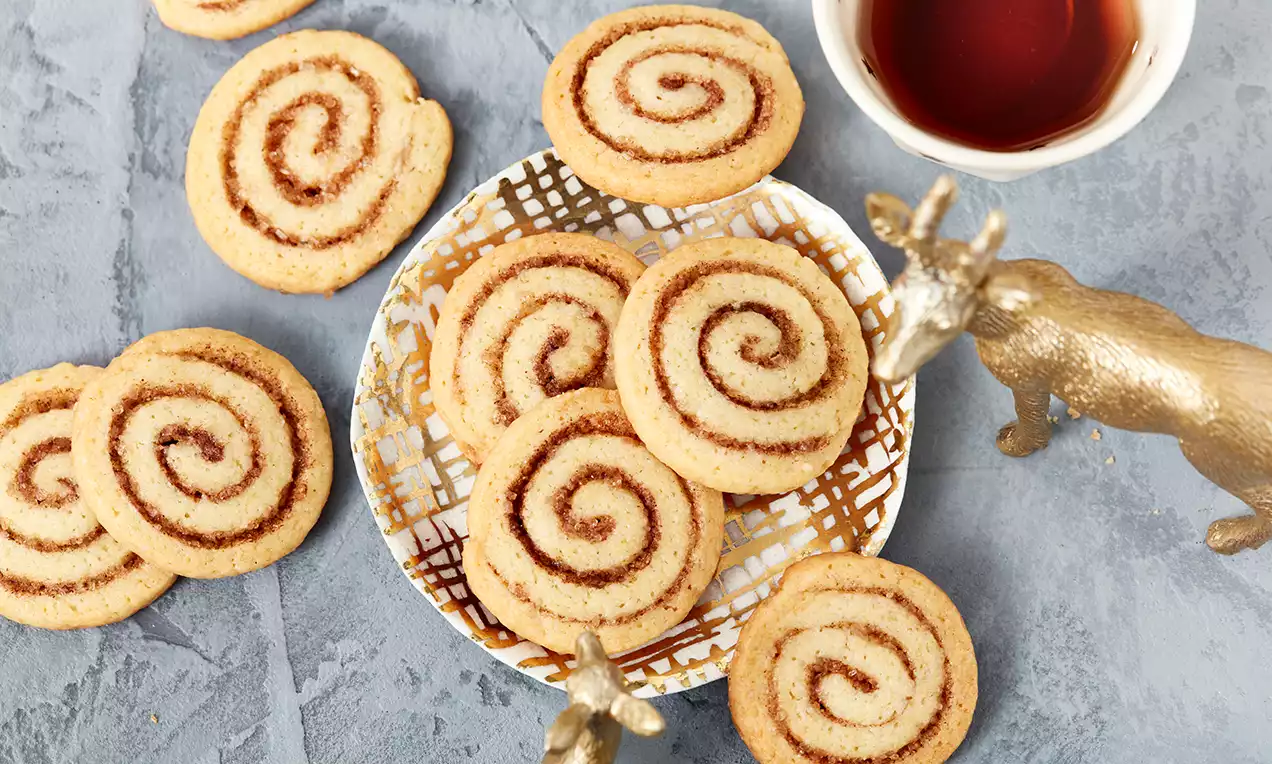 Hellbraune Zimtschnecken-Kekse mit spiralförmiger Füllung auf gemustertem Teller, daneben goldene Dekofiguren und Schale mit Tee