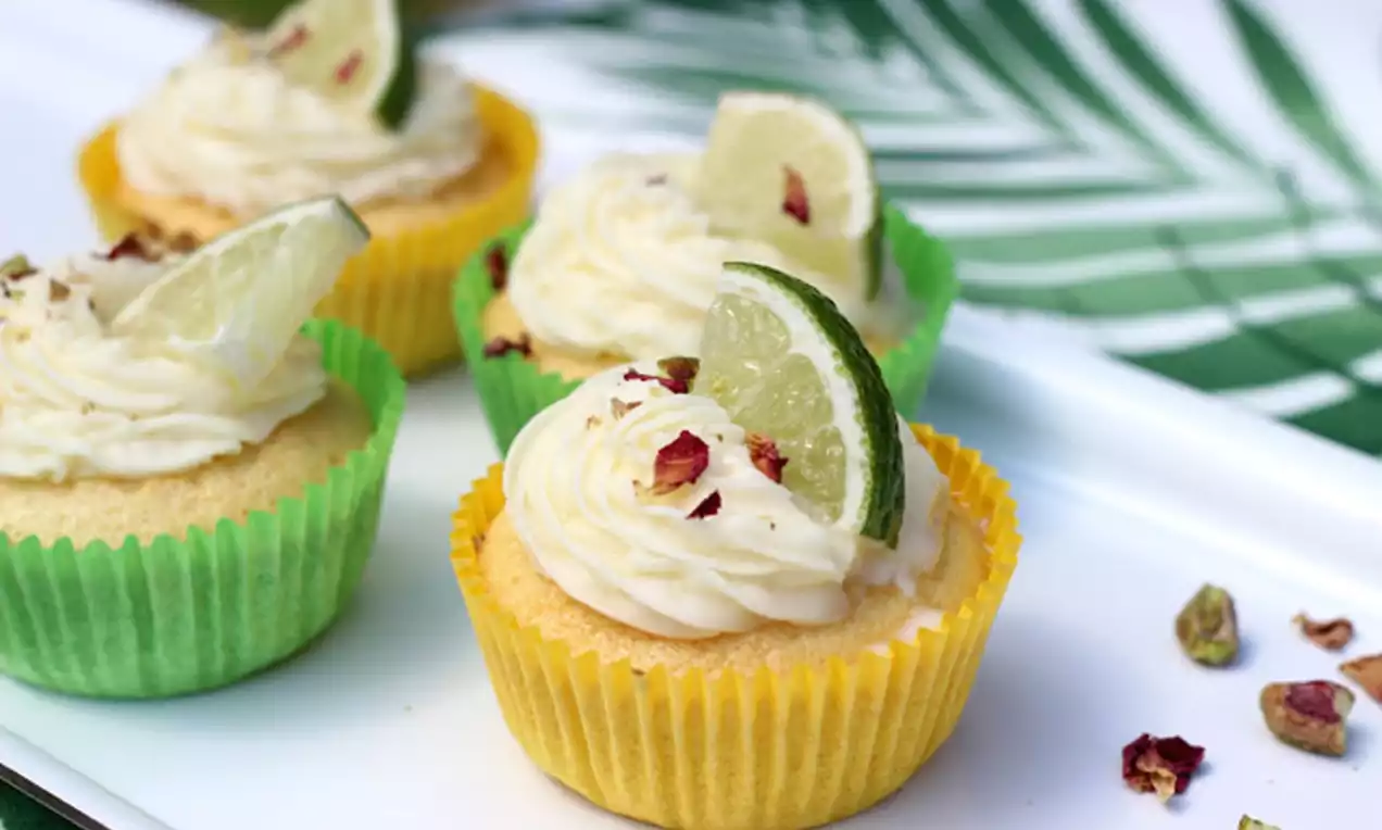 Cupcakes moelleux dans des caissettes jaunes et vertes, garnis de crème légère, quartiers de citron vert et éclats de pistache sur un plateau blanc