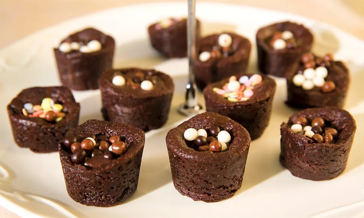 Малки цилиндрични шоколадови сладки с влажна текстура, гарнирани с цветни перли и шоколадови топчета, подредени върху бяла чиния