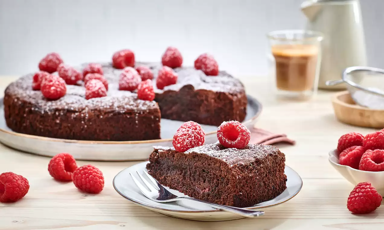 Schokotarte mit glänzend dunkler Oberfläche und frischen roten Himbeeren, auf einem Teller angerichtet, daneben Kaffee und weiteres Zubehör