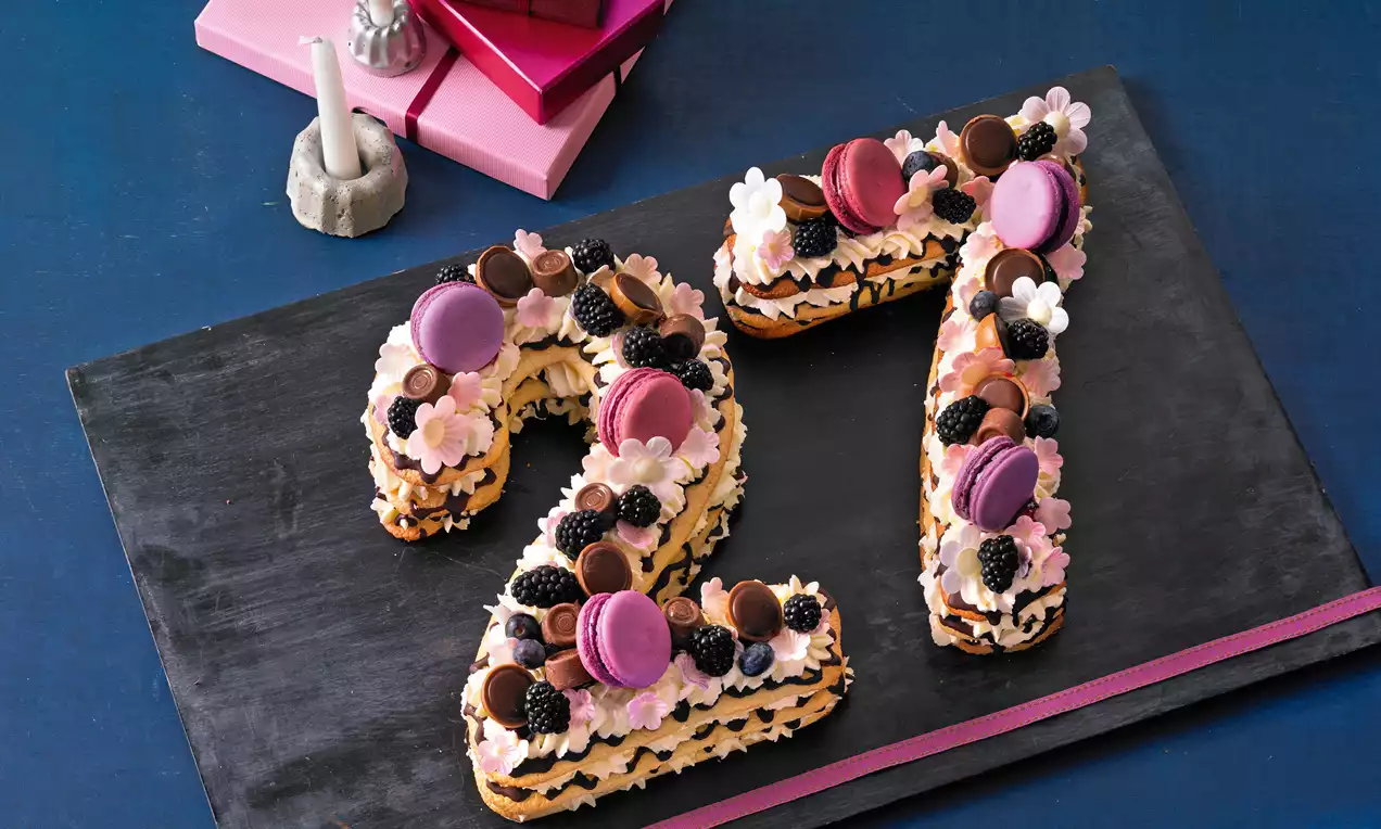 Zahl 27 aus hellbraunem Teig mit rosa-weißer Creme, dekoriert mit lila Macarons, Schokokugeln, Brombeeren und Blüten auf dunkler Platte