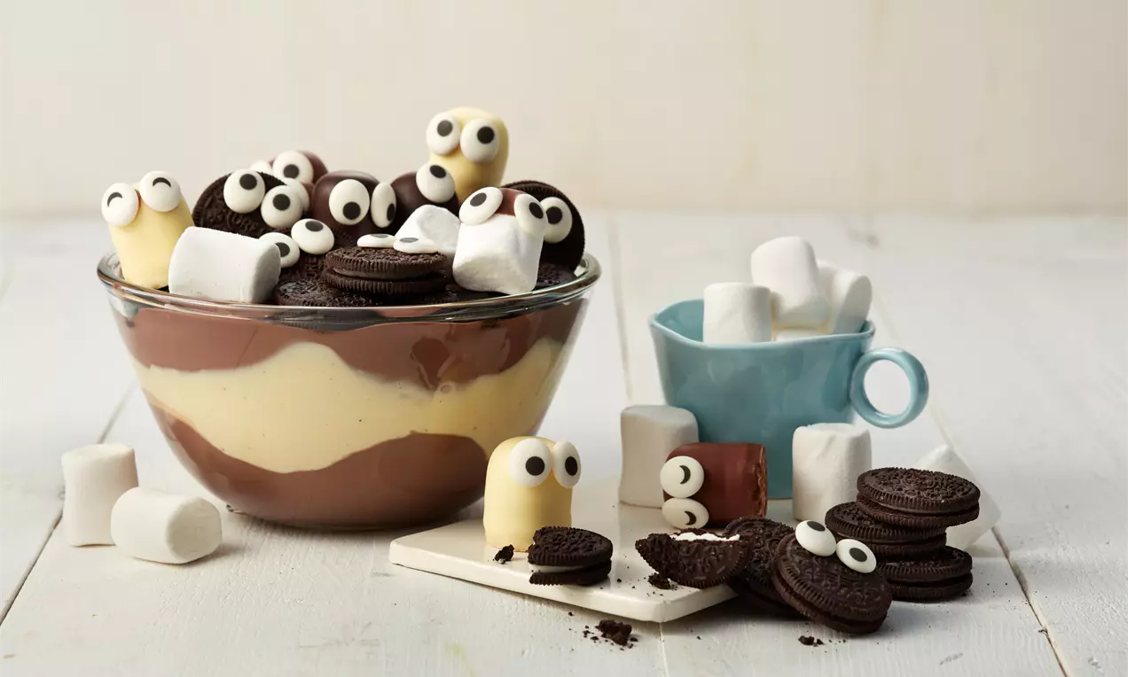 In einer Glasschale gestreifter Pudding mit gelber Vanille- und brauner Schokoschicht, dekoriert mit Oreos, Marshmallows und Zuckeraugen