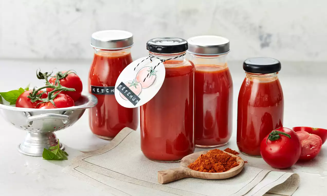 Glasflaschen mit rotem Gewürzketchup auf hellem Stoff, flankiert von frischen Tomaten in silberner Schale und rotem Gewürz auf Holzlöffel