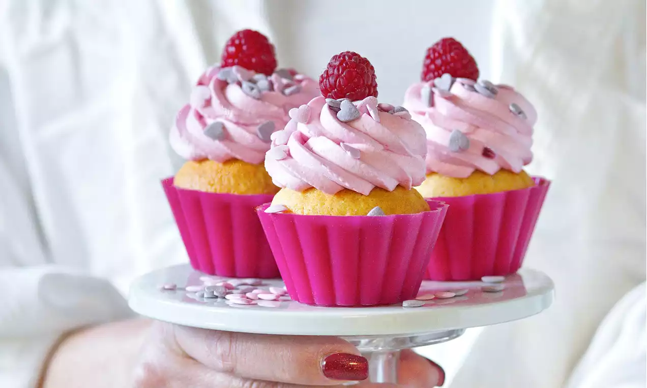 Tři světlé žluté cupcakes v růžových košíčcích, každý zdobený malinovou růžovou šlehačkou, čerstvou malinou a stříbrnými cukrovými srdíčky