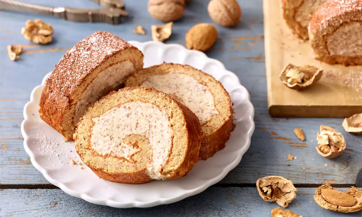 Feine Nussrolle mit goldbrauner Kruste und hellem Nusscremekern, in Scheiben geschnitten auf einem weißen Teller mit Walnussschalen