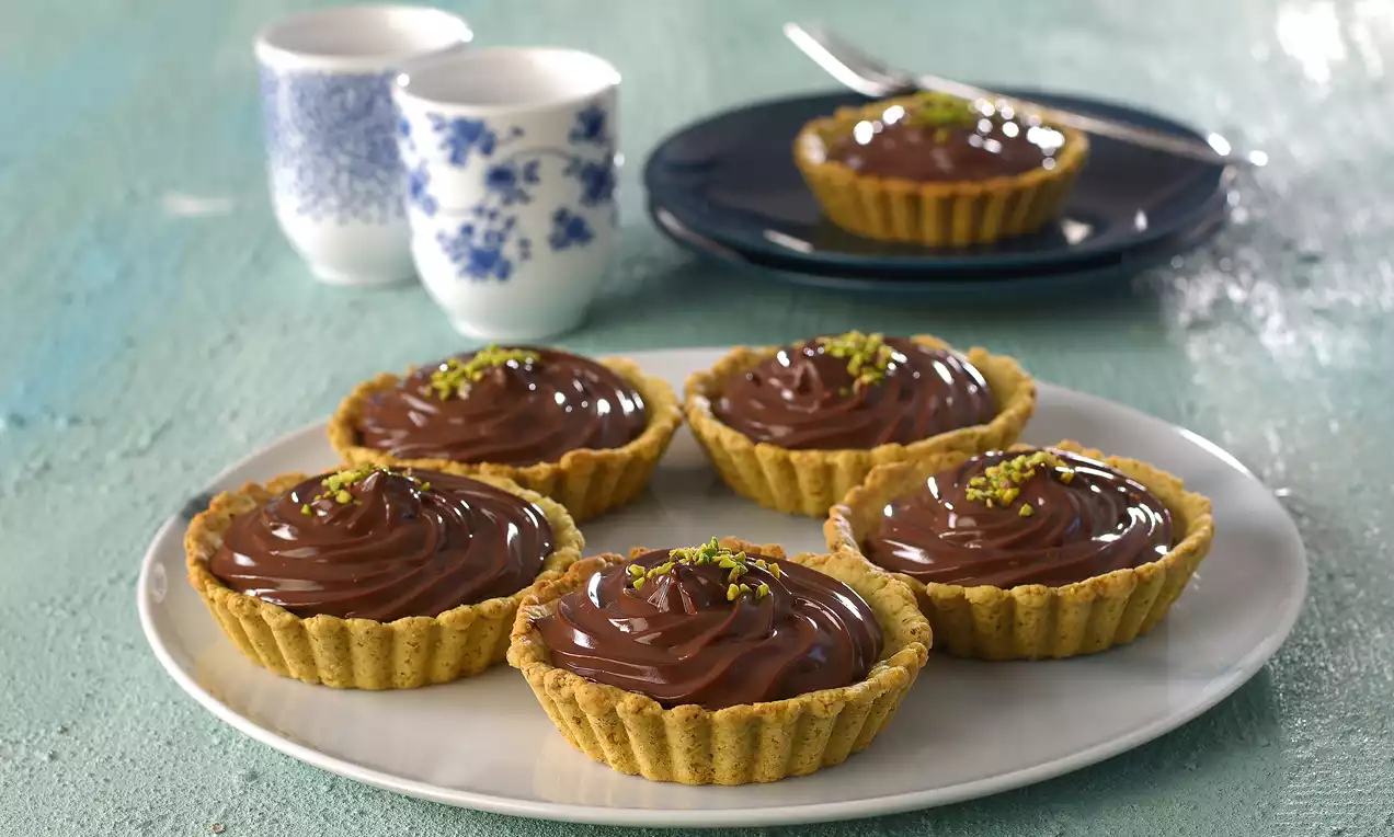 Kahverengi çıtır tabanlı tartoletler, kremamsı çikolata dolgusu ve üzerlerine serpilmiş ince kıyılmış antep fıstıkları ile beyaz tabakta