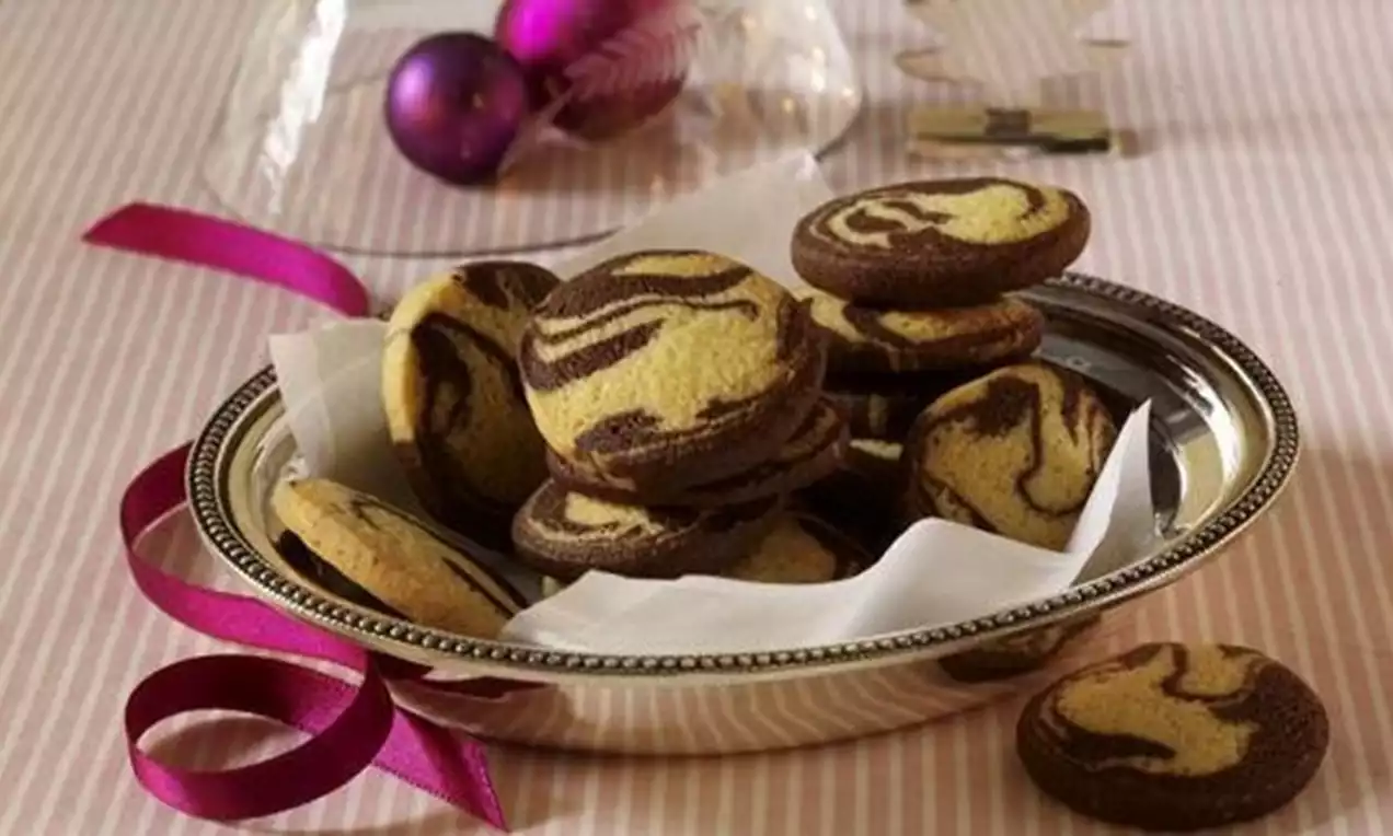Biscuits marbrés aux spirales brunes et dorées disposés sur du papier blanc dans une assiette argentée ornée d’un ruban rose