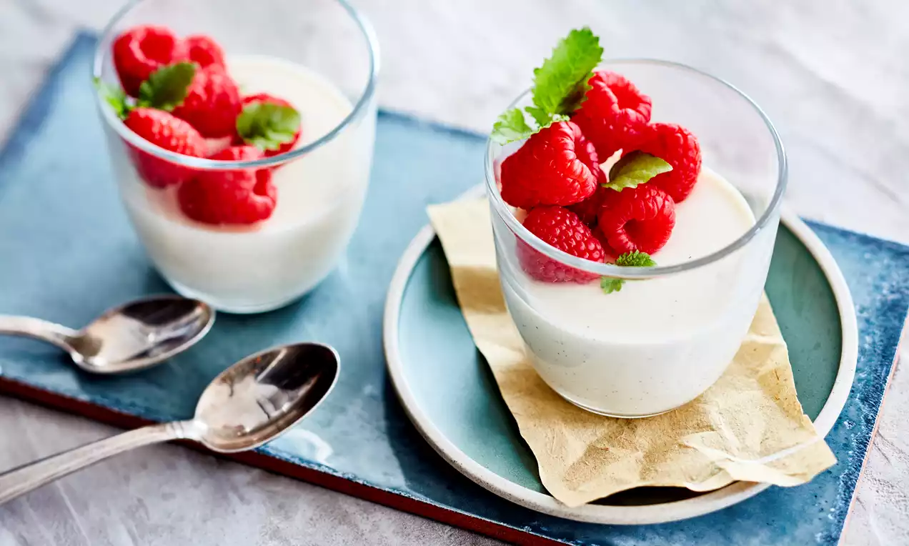 Panna cotta crémeuse couleur ivoire dans des verrines transparentes, surmontée de framboises rouges et de feuilles de menthe vertes