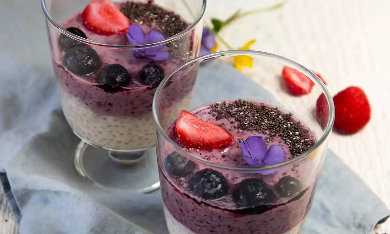 Ve sklenicích vrstvy světlé krémové báze a sytě fialového borůvkového smoothie, ozdobené chia semínky, lesním ovocem a fialovými kvítky