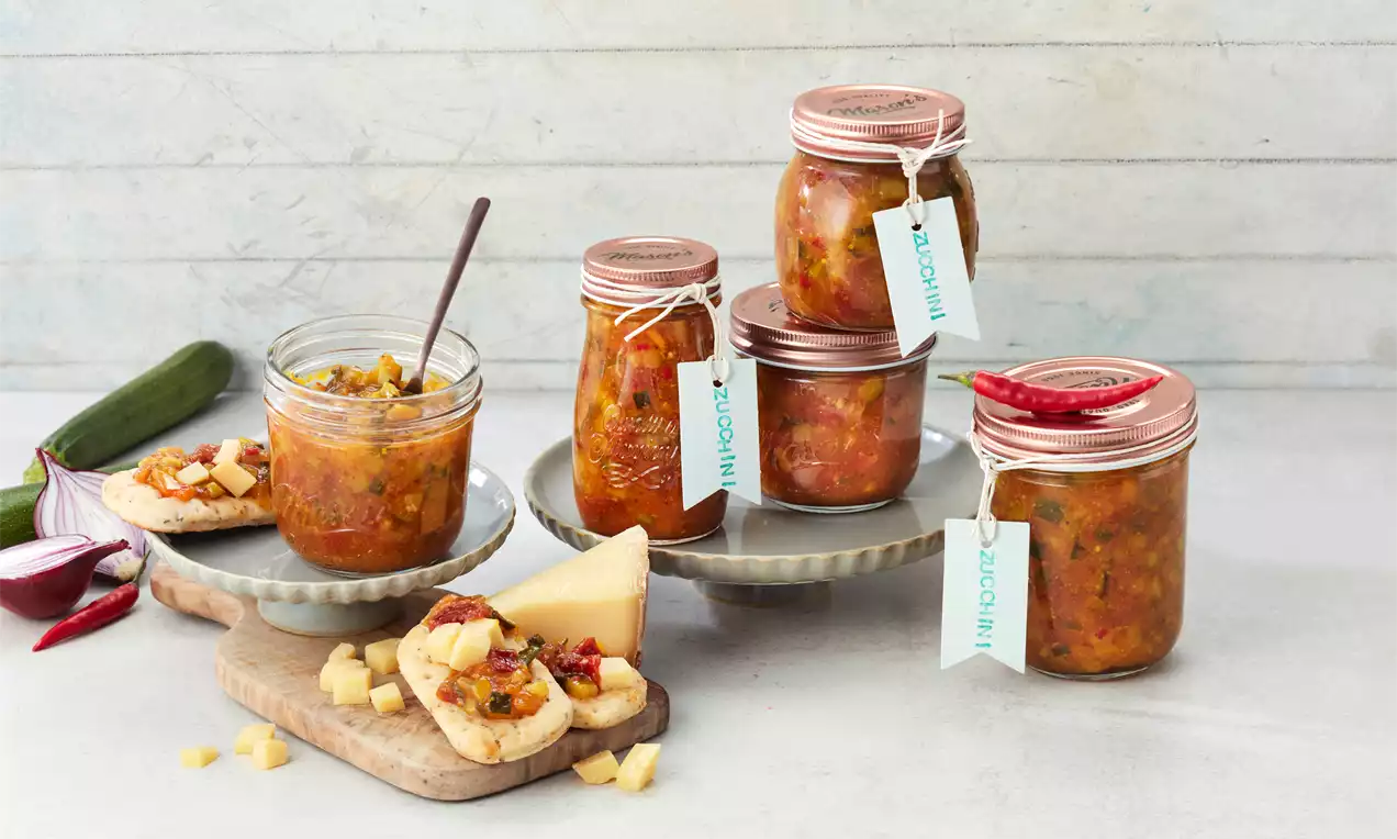 Gläser mit orangem Zucchini-Chutney, teils geöffnet, mit roten Deckeln und Etiketten auf Tellern, serviert neben Brotscheiben mit Käse