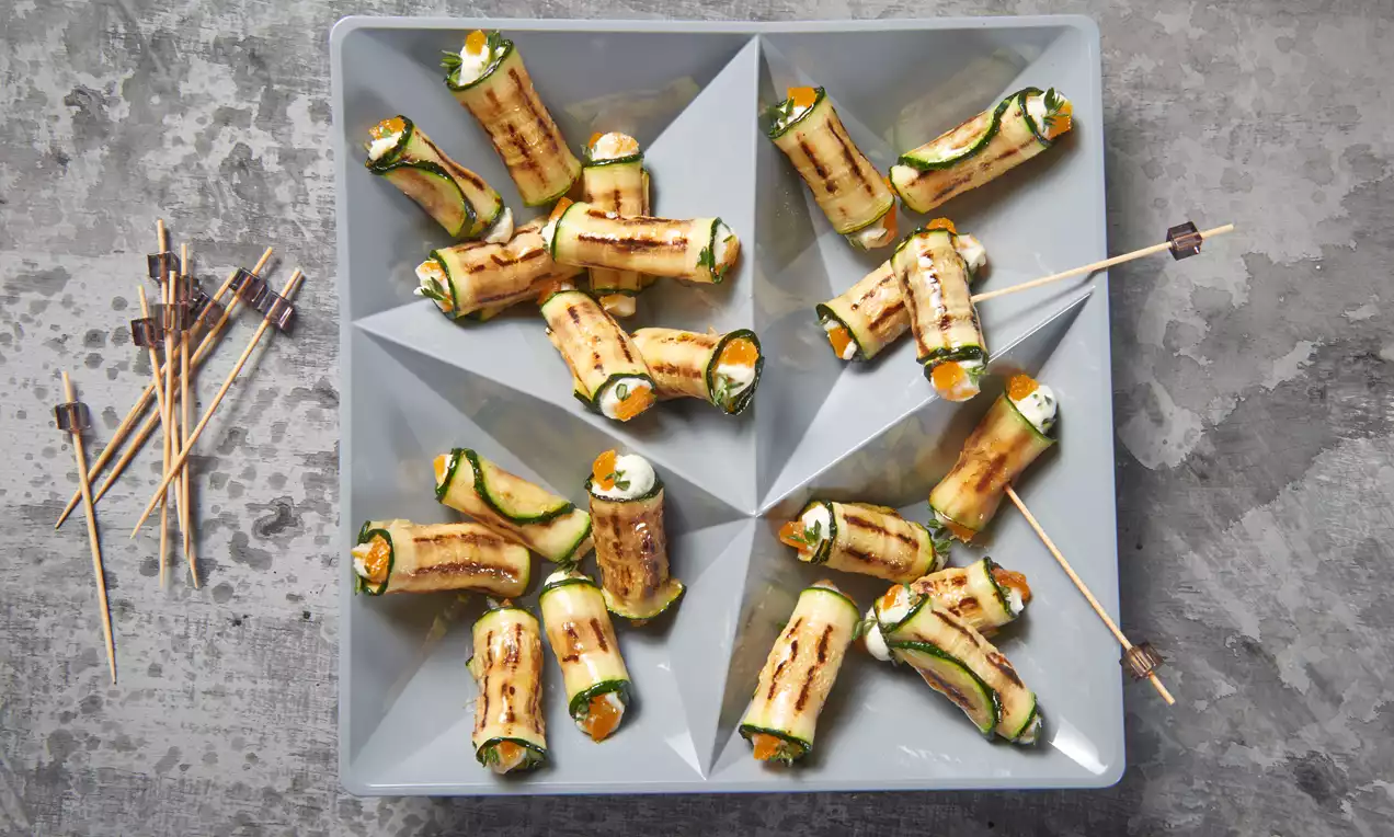 Gegrillte Zucchiniröllchen mit cremiger Frischkäsefüllung und orangefarbenem Gemüse, dekorativ auf grauer Servierplatte angerichtet
