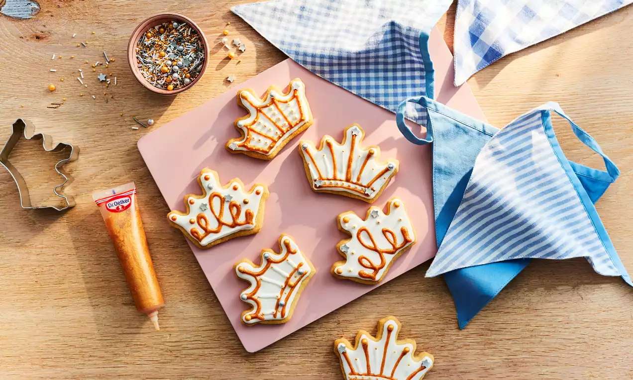 Golden crown-shaped biscuits with intricate white icing details and caramel accents arranged on a pink board with decorative items nearby