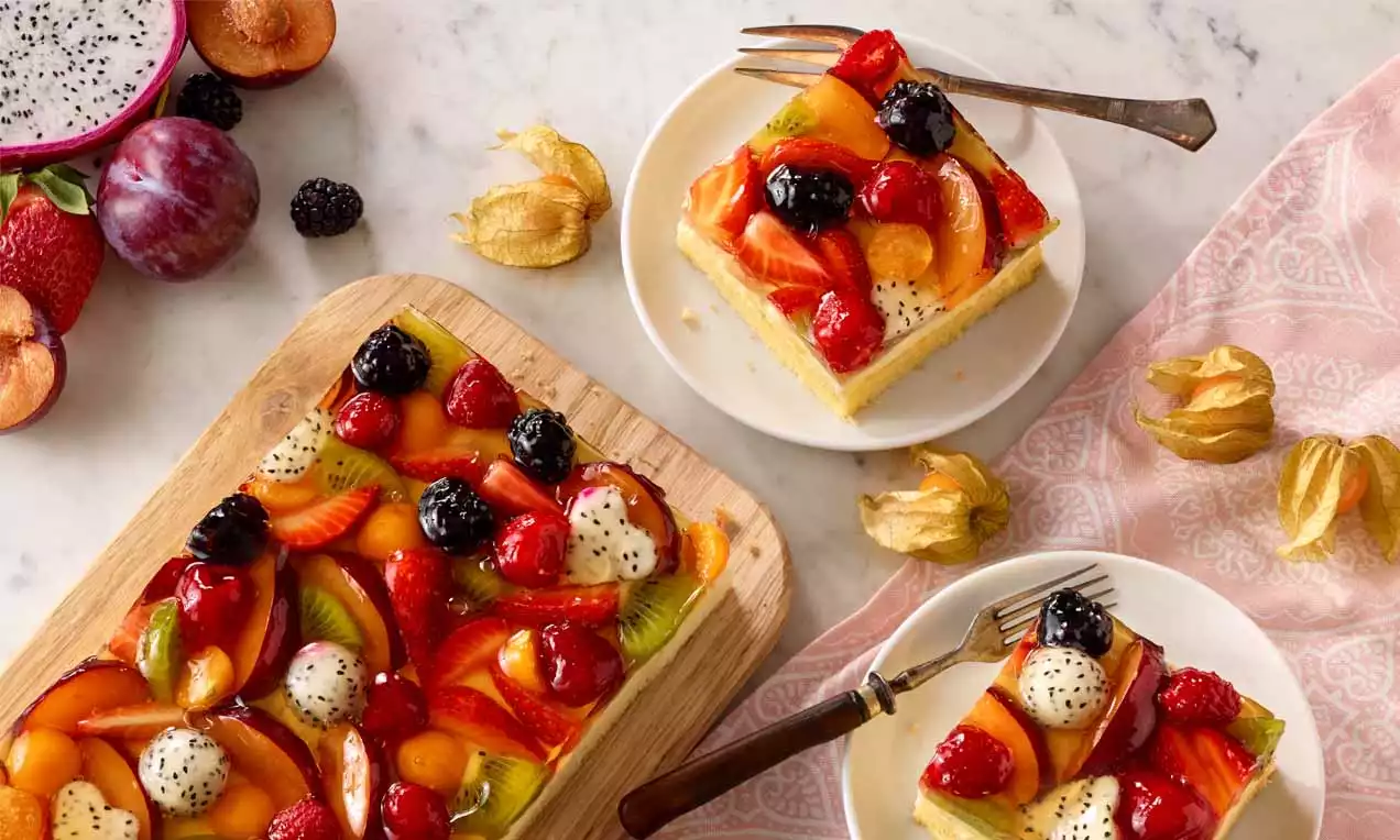 Bunte Obst-Pudding-Schnitten auf hellem Holzbrett und weißen Tellern, belegt mit glänzenden Früchten wie Kiwi, Beeren und Physalis