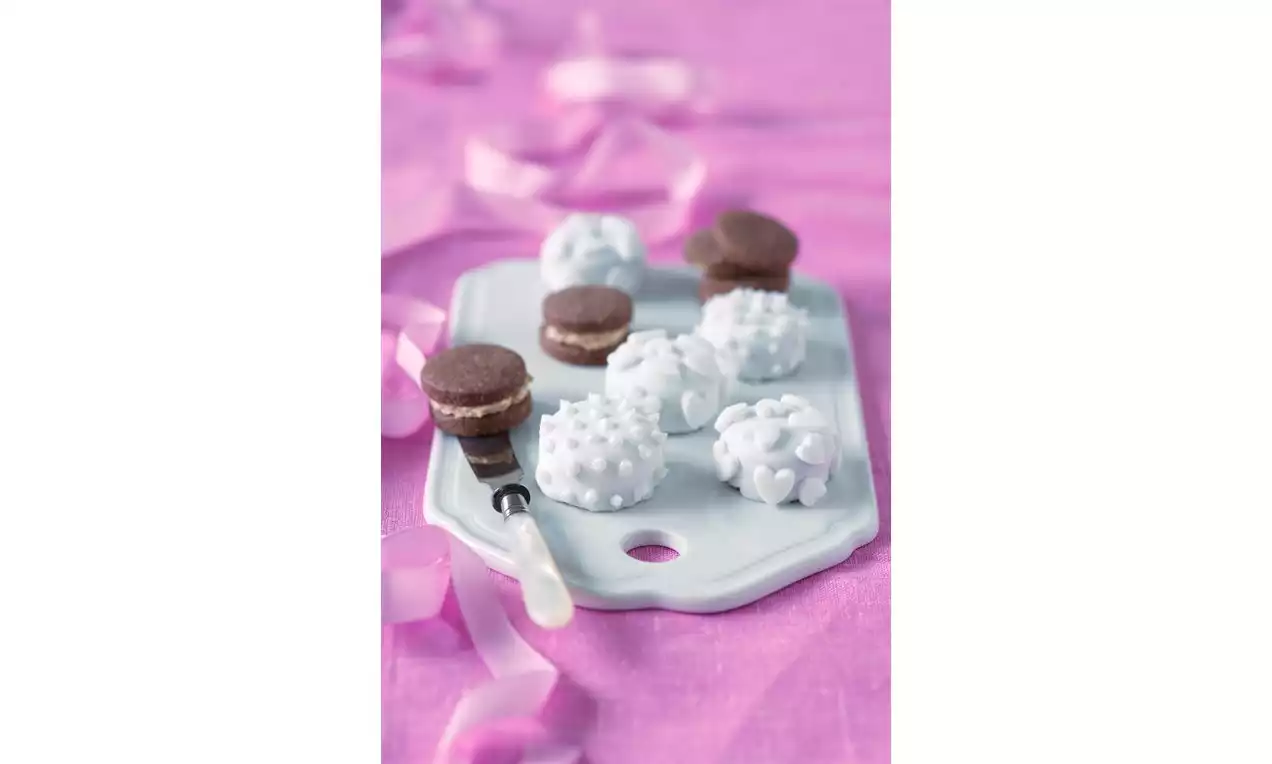 Čokoládové sušienky plnené krémom a biele cukrové guľky s bodkami na bielom podnose, zdobené ružovou stuhou na textilnom podklade