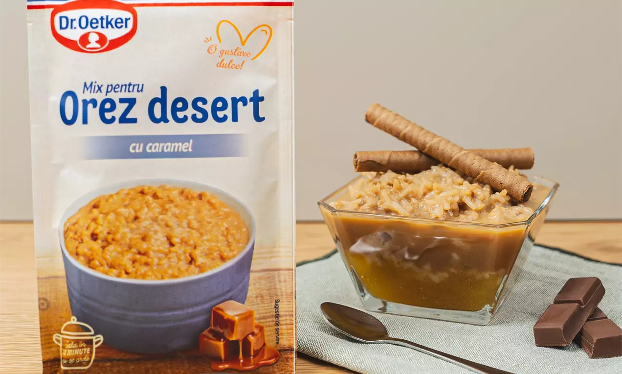 Orez cremos de culoare caramel, așezat într-un bol de sticlă transparent, decorat cu rulouri crocante și servit pe un șervet gri