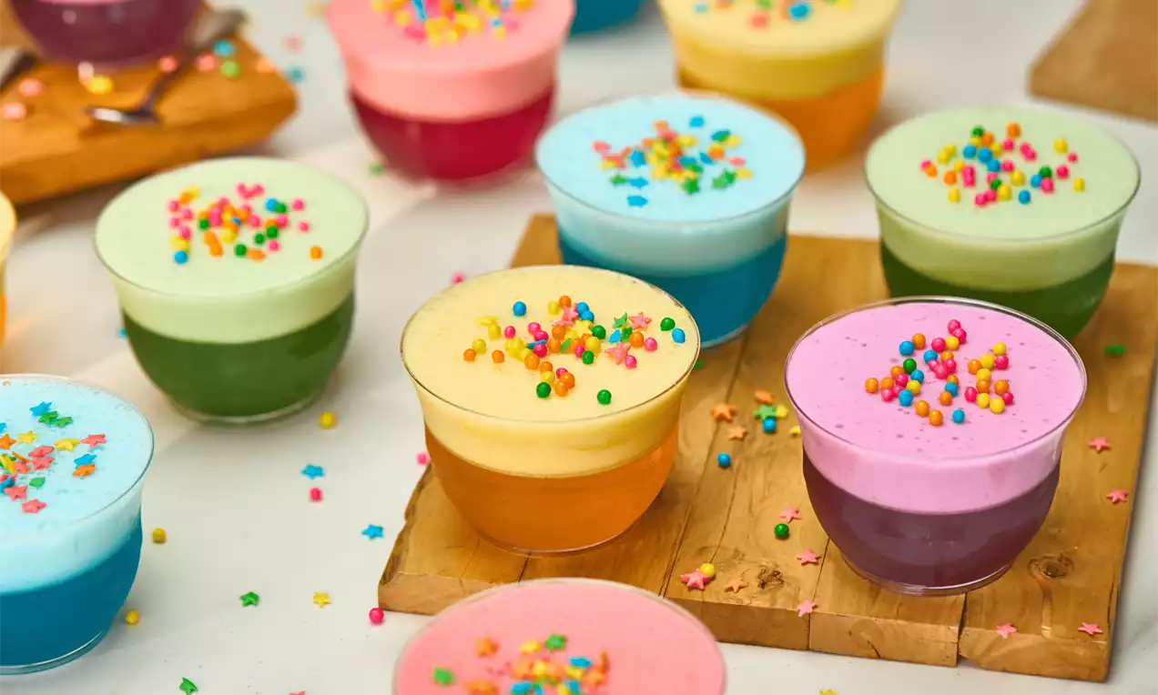 Copos transparentes com camadas vibrantes de gelatina colorida cobertas por creme suave decorado com confeitos multicoloridos sobre tábua de madeira