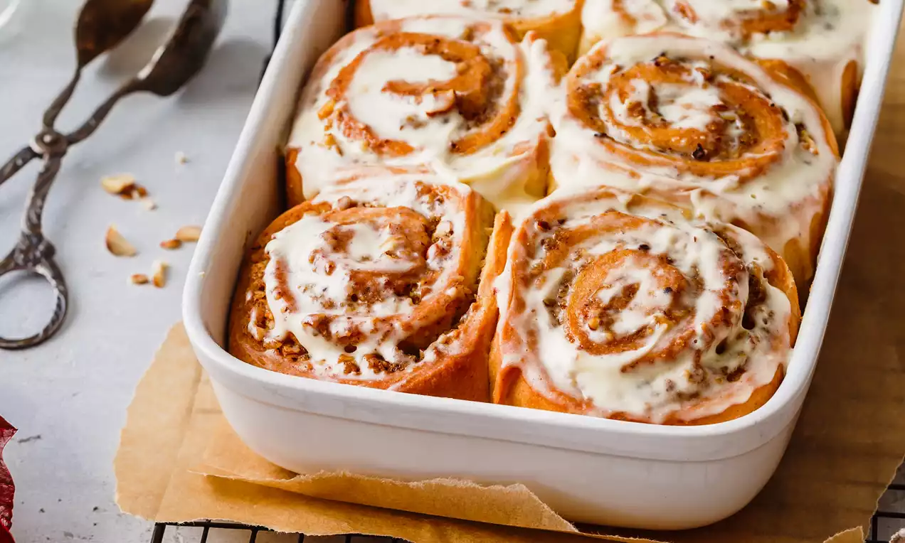 Picture - Soft, golden-brown cinnamon rolls swirled with nuts, topped with creamy white vanilla frosting, arranged in a white baking dish