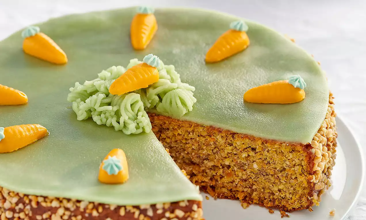 Torta ricoperta di marzapane verde e decorata con carotine arancioni modellate, su base dorata con bordo di granella di nocciole