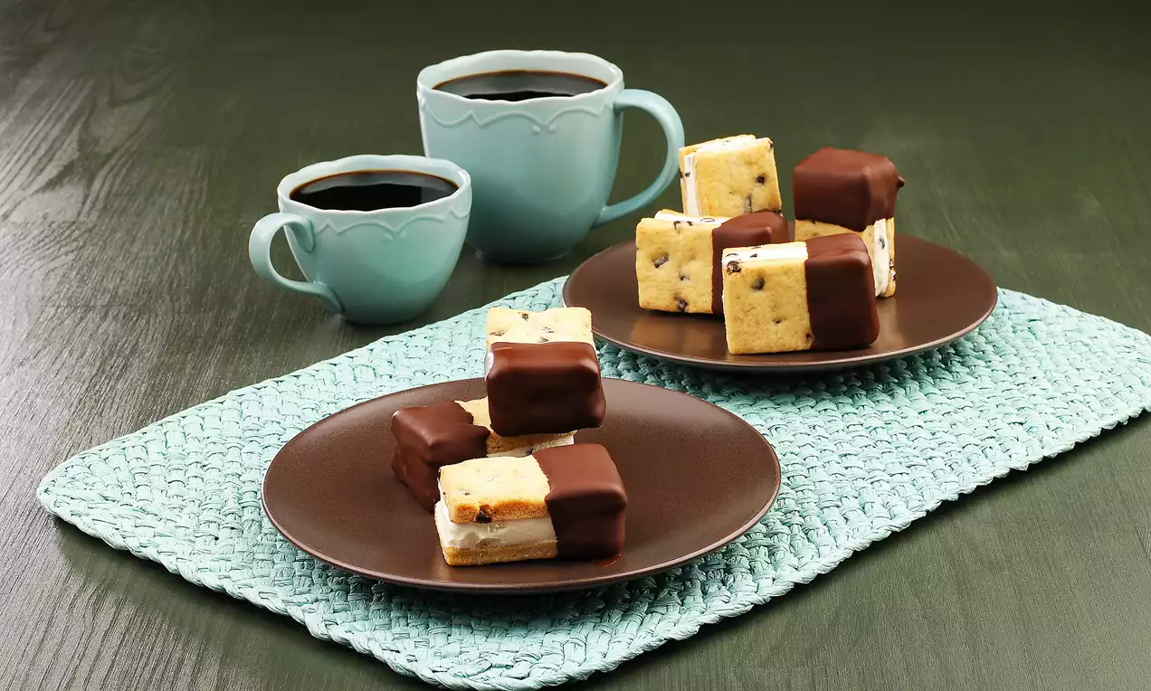 Kenarları çikolataya batırılmış, kare şeklinde kremalı ve çikolata parçalı dilimler kahverengi tabakta, arka planda iki porselen fincan.