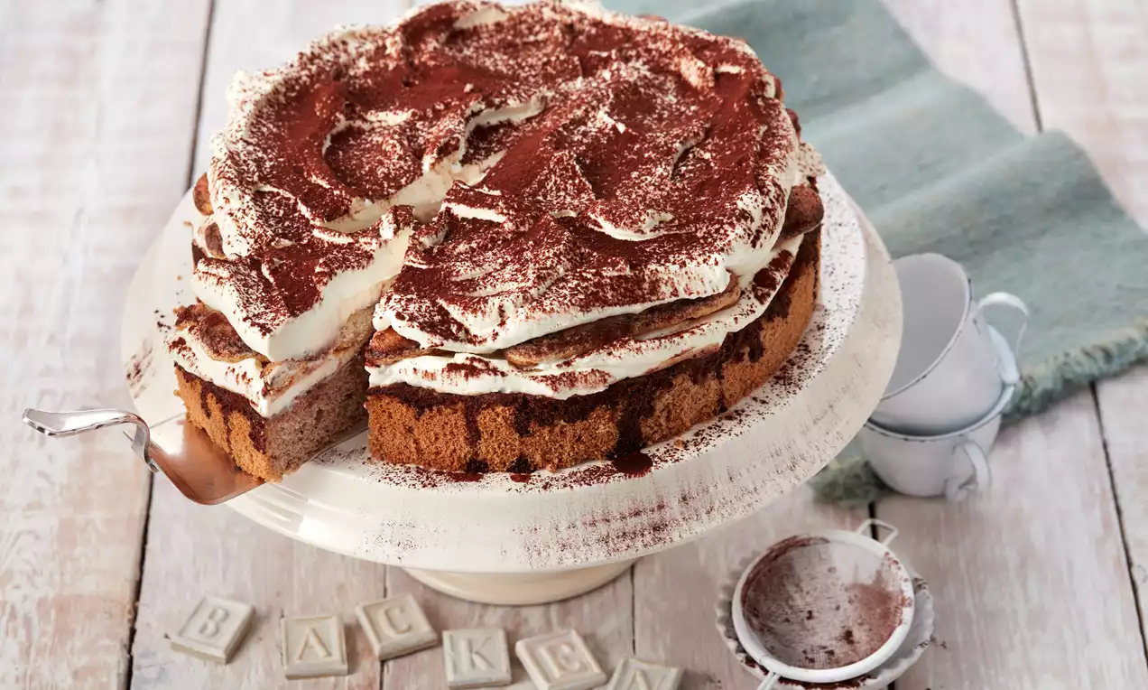 Tiramisu-Torte mit lockerem Löffelbiskuit, geschichteter Creme und Kakao, auf weißer Tortenplatte mit Sieb und Buchstaben-Dekoration drapiert