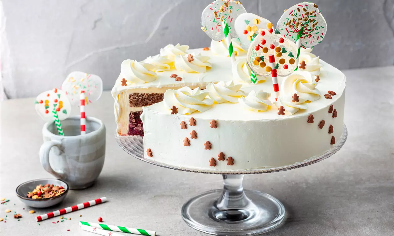 Сметанова торта със снежнобял крем, украсена с цветни близалки и поръсена с малки шоколадови фигурки върху прозрачна поставка