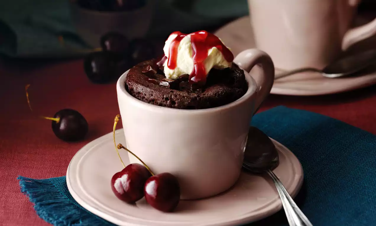 Rich dark chocolate mug cake topped with cream and red cherry sauce, served in a pale pink mug on a matching saucer with fresh cherries