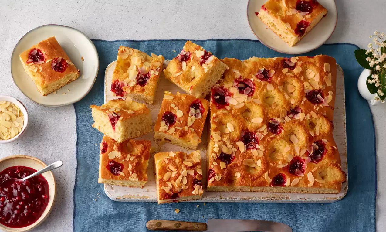 Goldbrauner Marzipan-Preiselbeer-Kuchen mit glänzenden Preiselbeeren und gerösteten Mandelblättchen, in Stücke geschnitten auf blauem Tuch