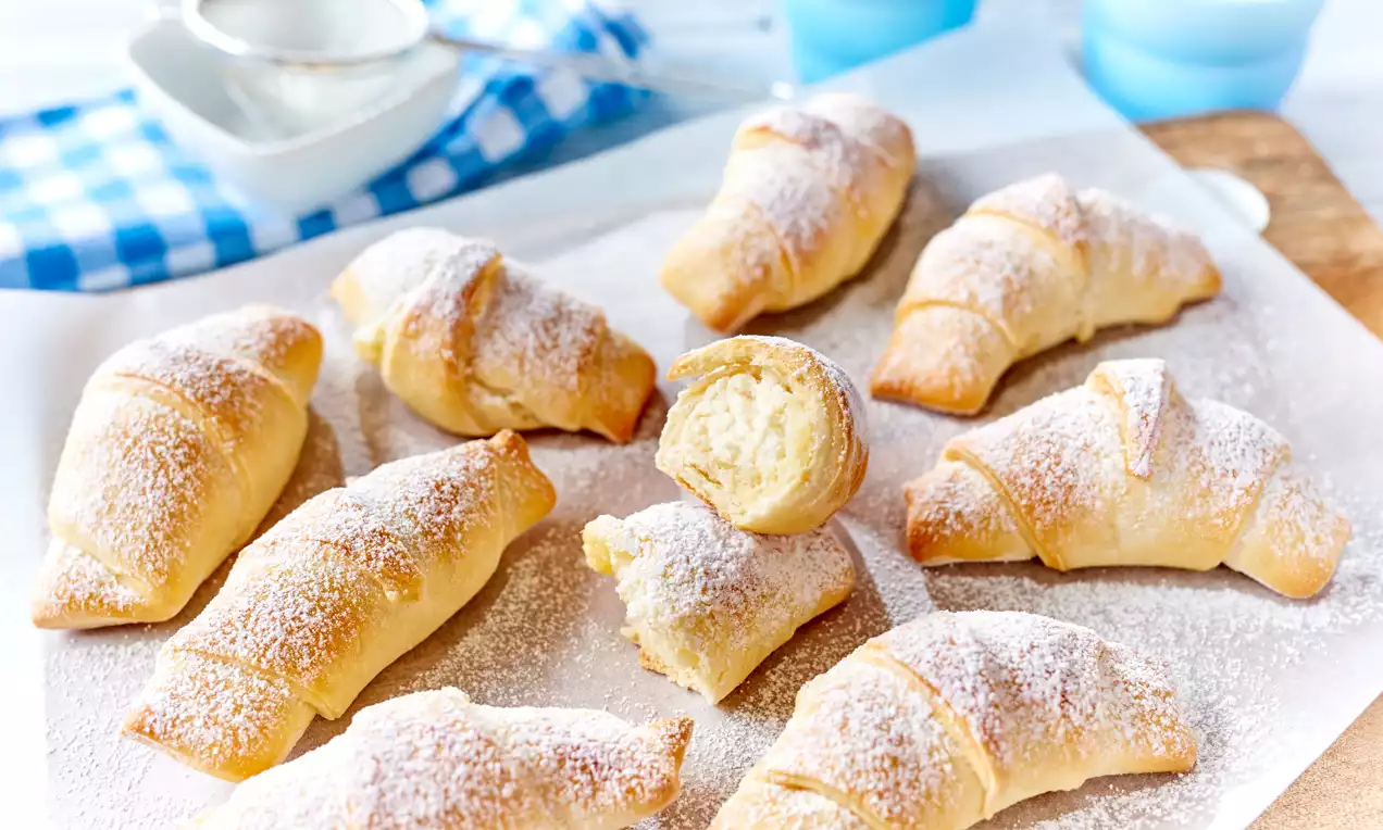 Goldbraune, leicht gebogene Topfenkipferln mit feinem Puderzucker bestäubt, auf weißem Backpapier locker angeordnet, ein Kipferl angeschnitten